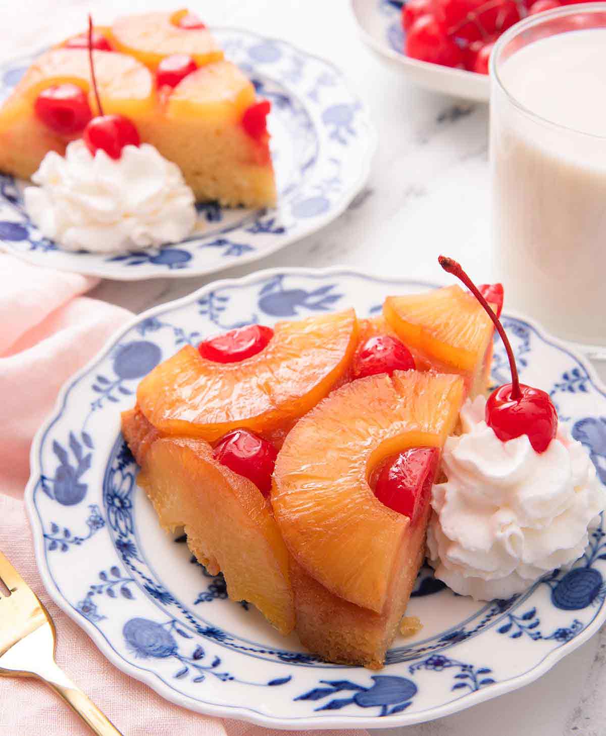 Two plates of pineapple upside down cake with one in the foreground, both with a dollop of whipped cream with a cherry on top.