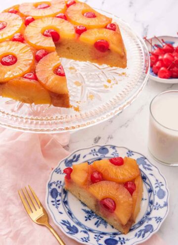 A pineapple upside down cake on a cake stand with a slice cut out and placed on a plate in front of it.