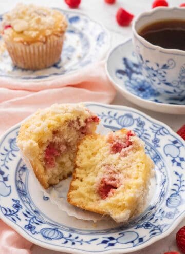A raspberry muffin cut in half on a blue and white plate beside a cup of coffee and another muffin in the back.