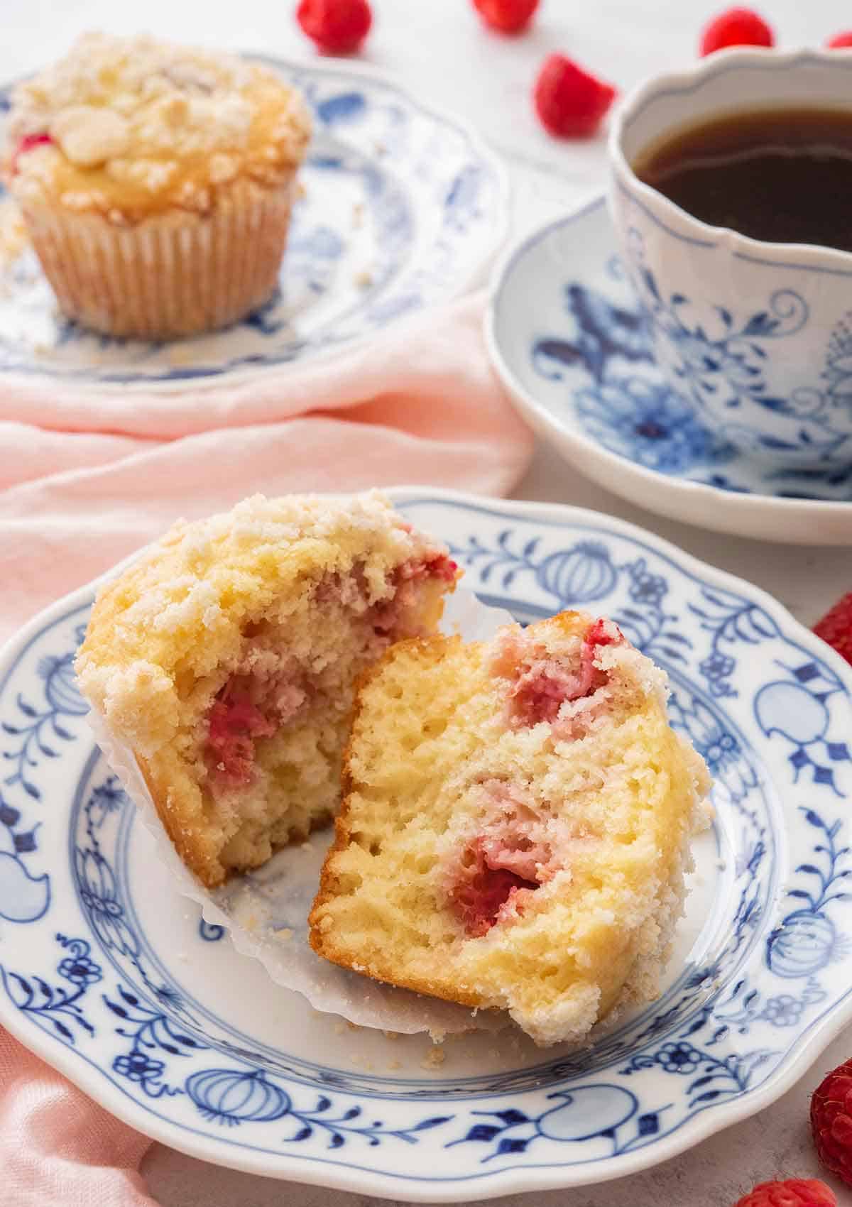 A raspberry muffin cut in half on a blue and white plate beside a cup of coffee and another muffin in the back.
