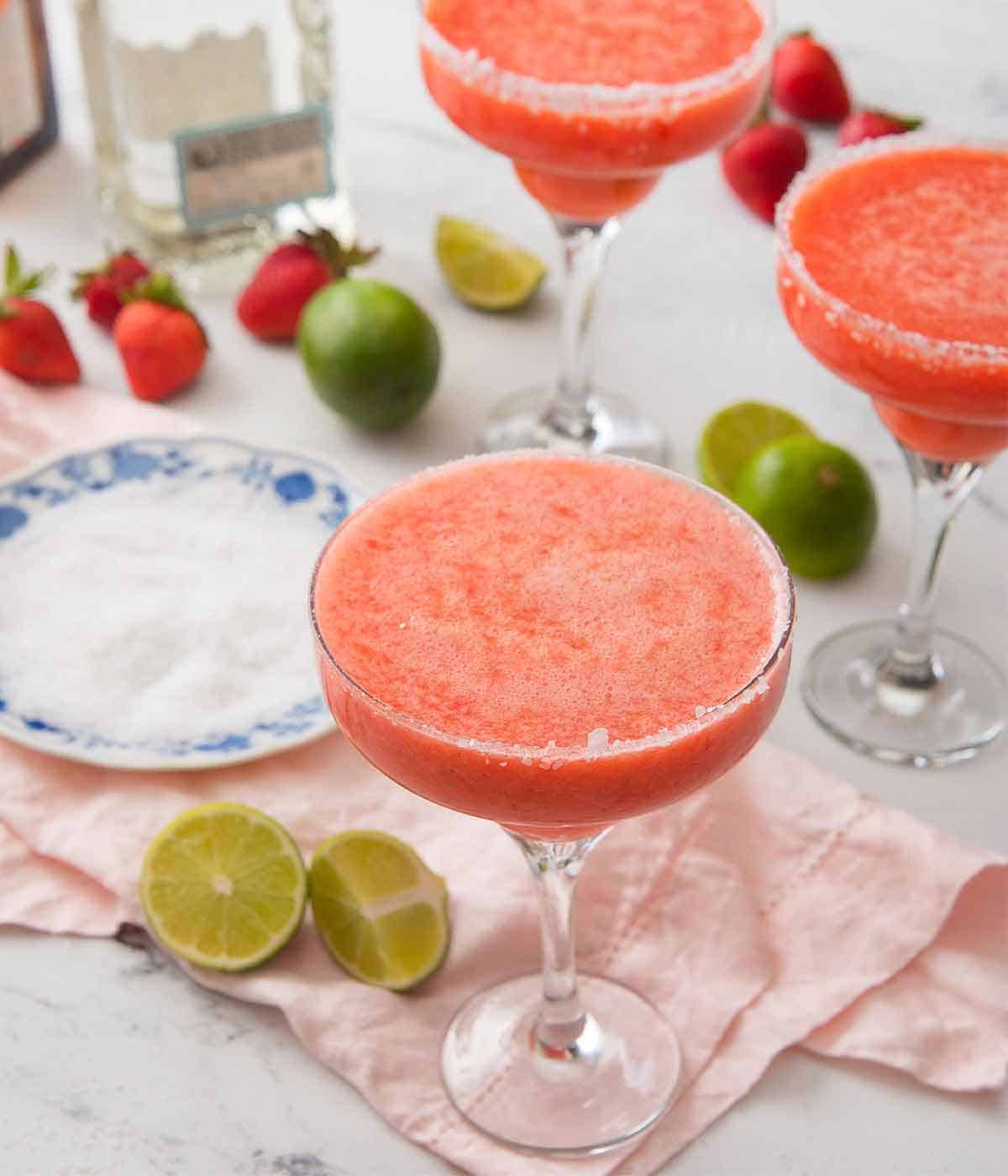 A set of three cocktail glasses with strawberry margarita beside a plate of salt, cut limes, and strawberries.