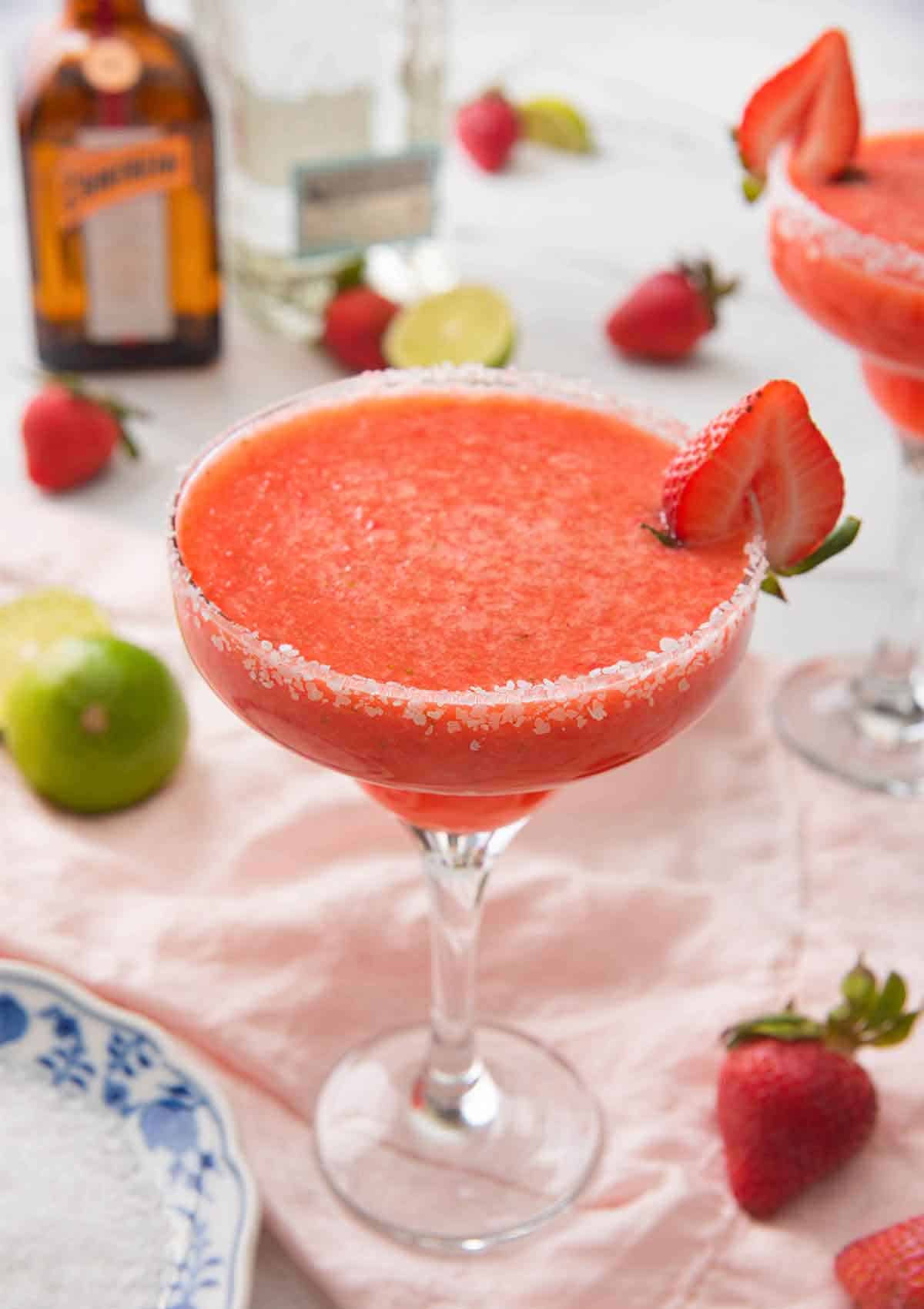 A strawberry margarita in a cocktail glass with a halved strawberry on the rim with the ingredients out of focus in the background.
