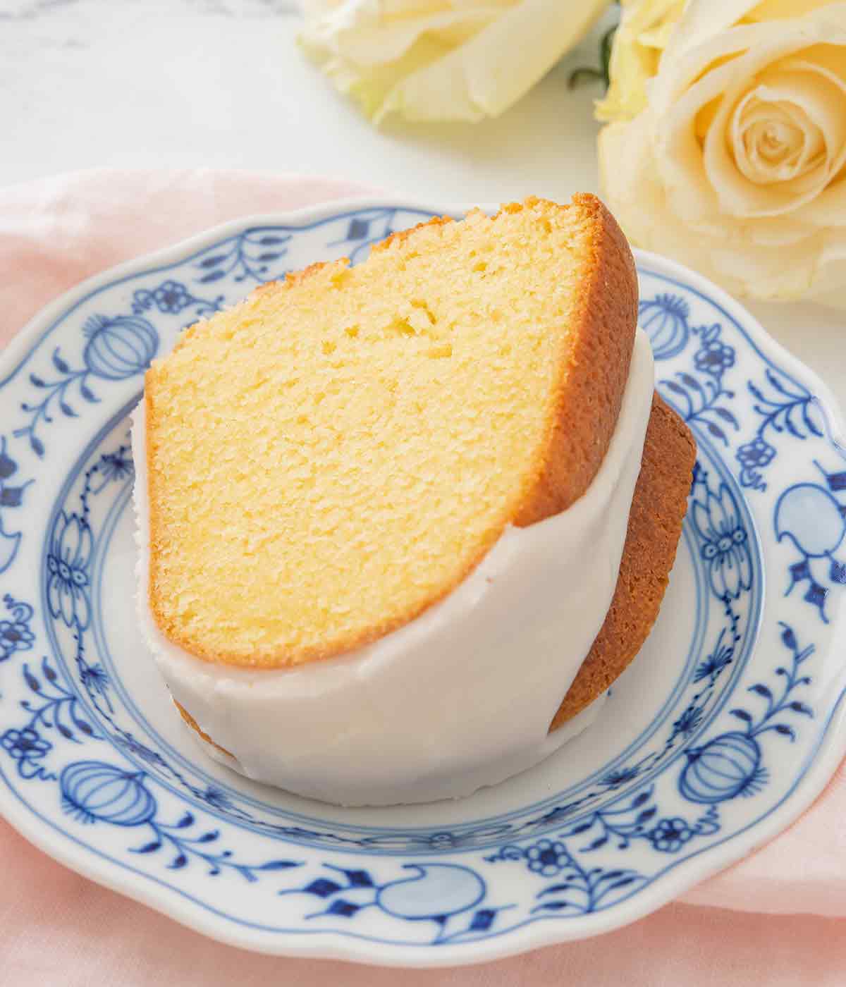 A slice of vanilla Bundt cake with glaze on a blue and white plate with white roses in the background.