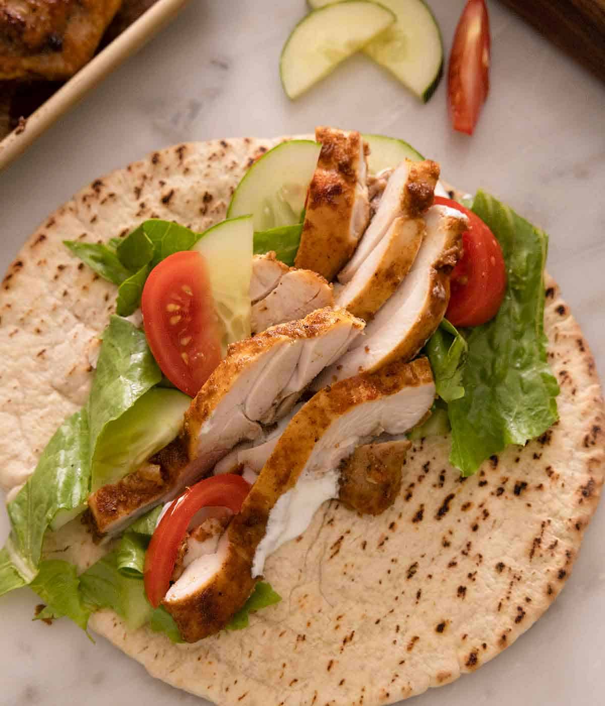 An opened pita with chicken shawarma with lettuce, tomatoes, and cucumbers.