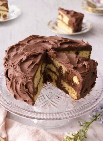 A cake stand with a chocolate frosted marble cake, with a quarter cut out. One cut slice is still on the stand and the other two are in the background on a plate.