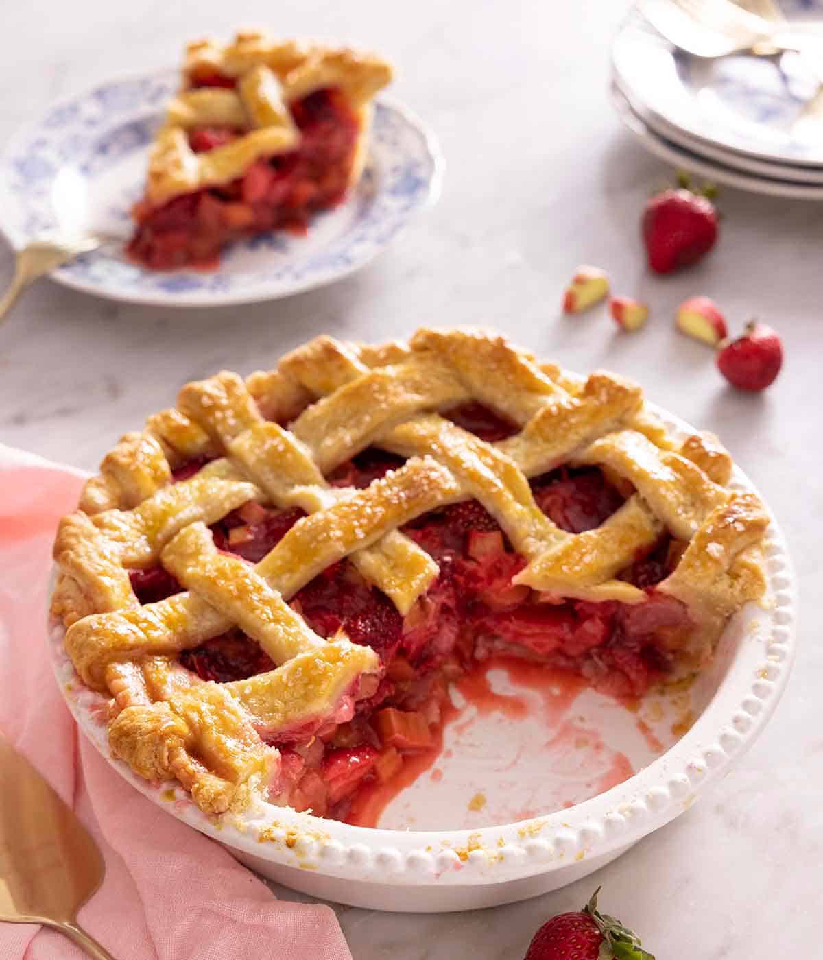 A white pie plate containing a strawberry rhubarb pie with a slice cut on, sitting on a plate in the background.