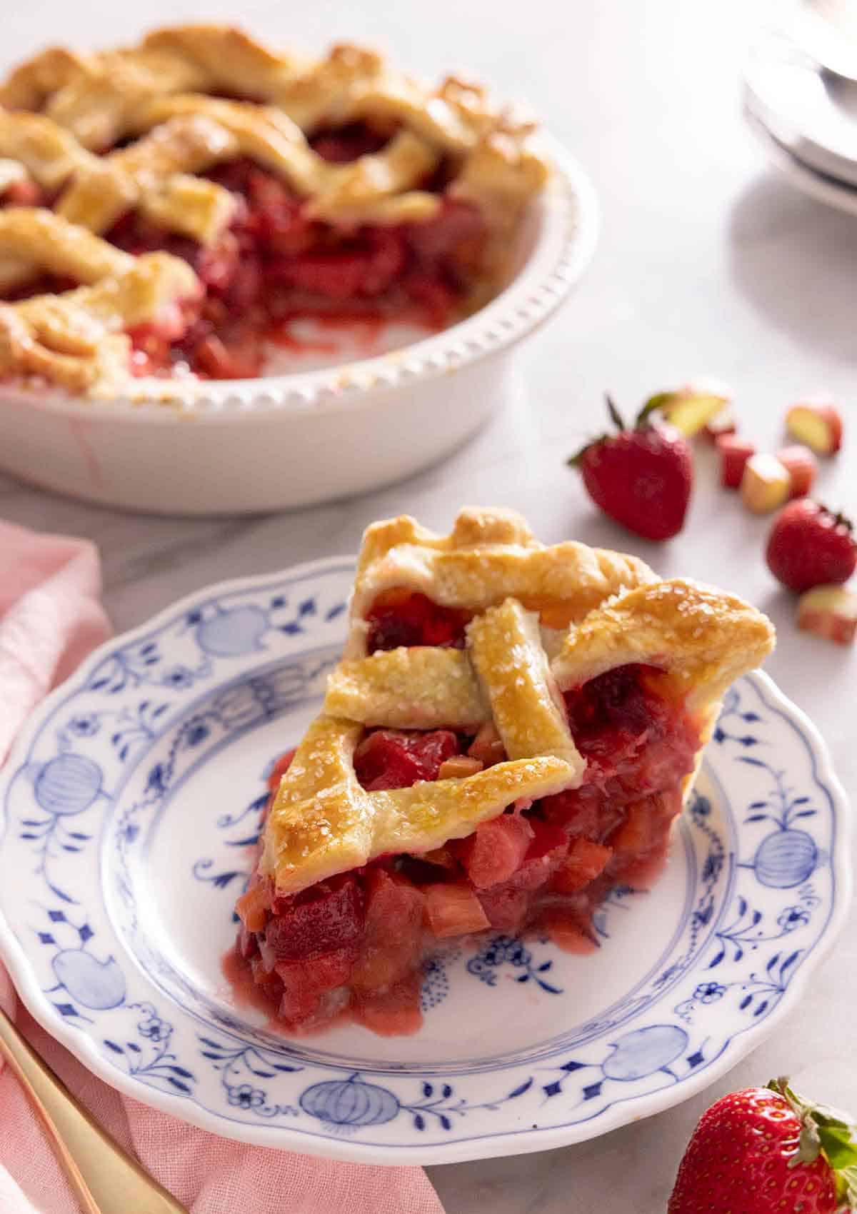 A slice of strawberry rhubarb pie on a plate with the rest of the pie in the ground out, out of focus.