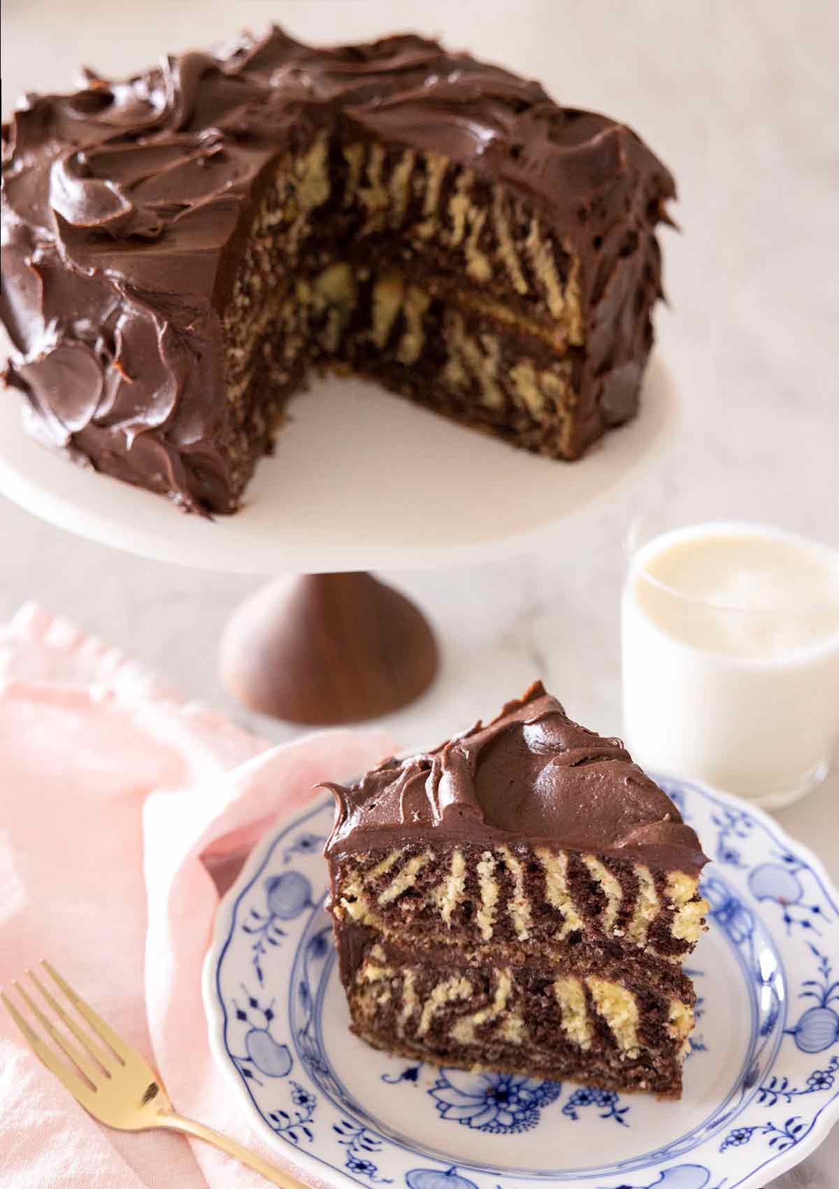 A slice of zebra cake on a plate in front of the rest of the cake behind it on a cake stand.