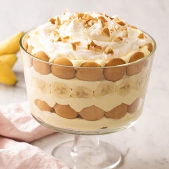 Banana pudding in a trifle dish with three bananas in the background. Pink napkin in front.