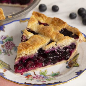 A piece of blueberry pie with a lattice top on a white plate next to some blueberries.