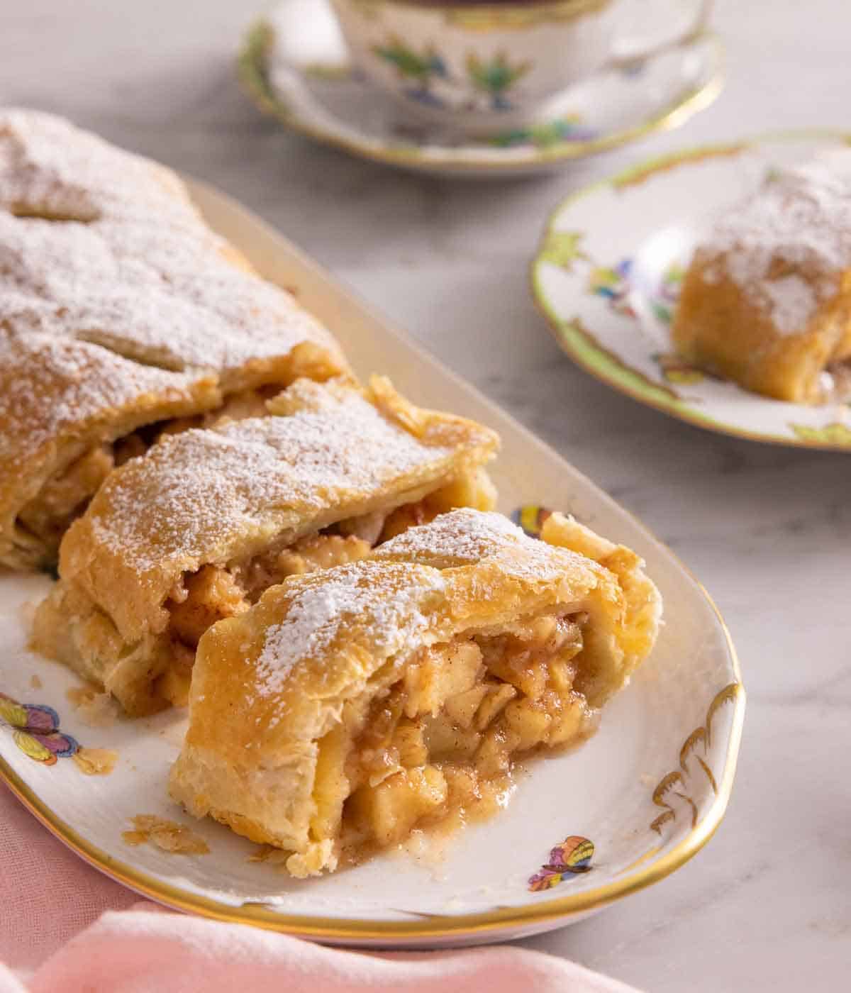 A platter with an apple strudel with two slices cut with a plate with a serving in the background.
