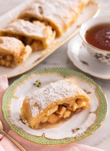 A serving of apple strudel on a plate in front of a cup of tea and more sliced apple strudel pieces.