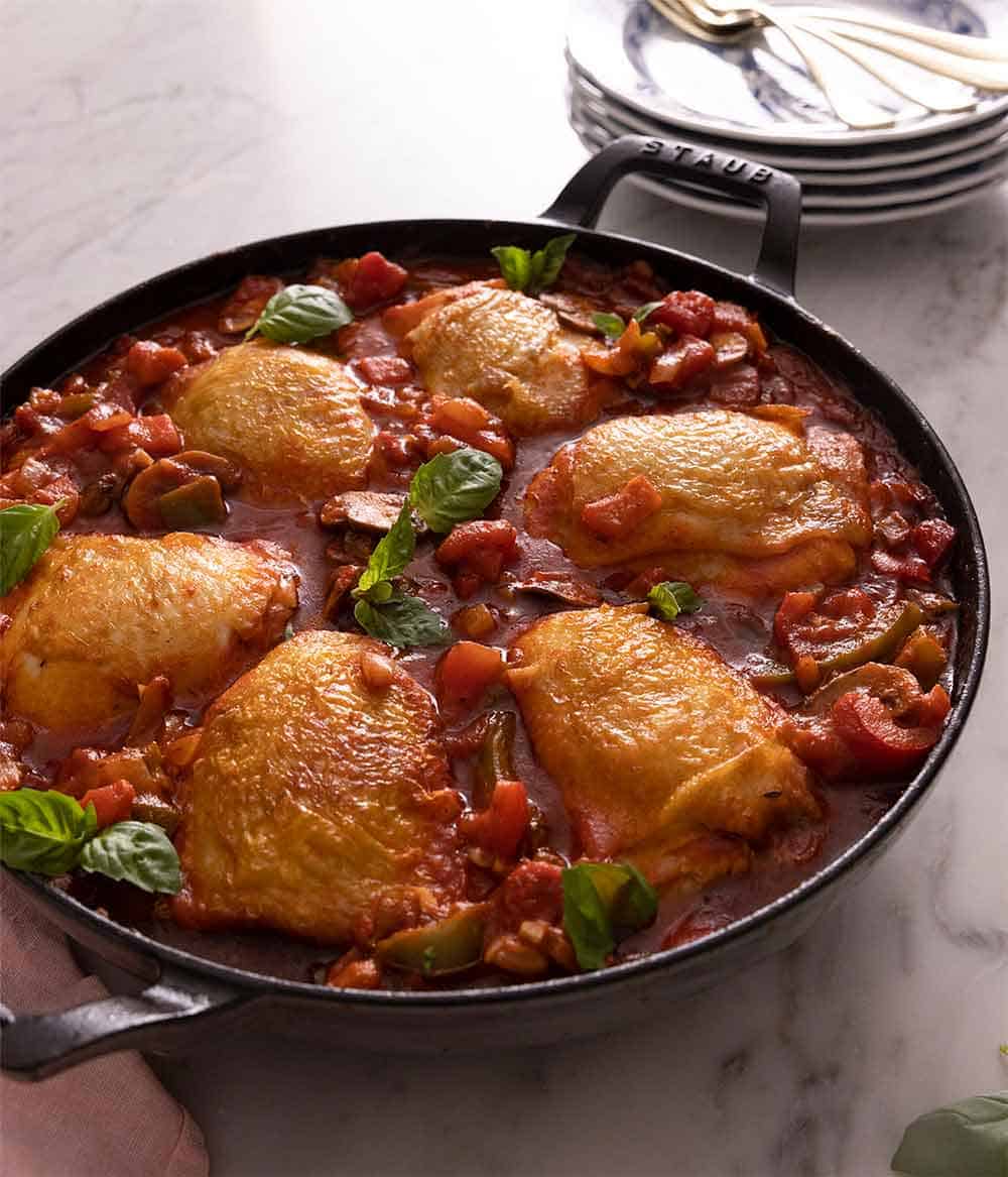 A large cast iron pan containing chicken cacciatore beside a stack of plates.