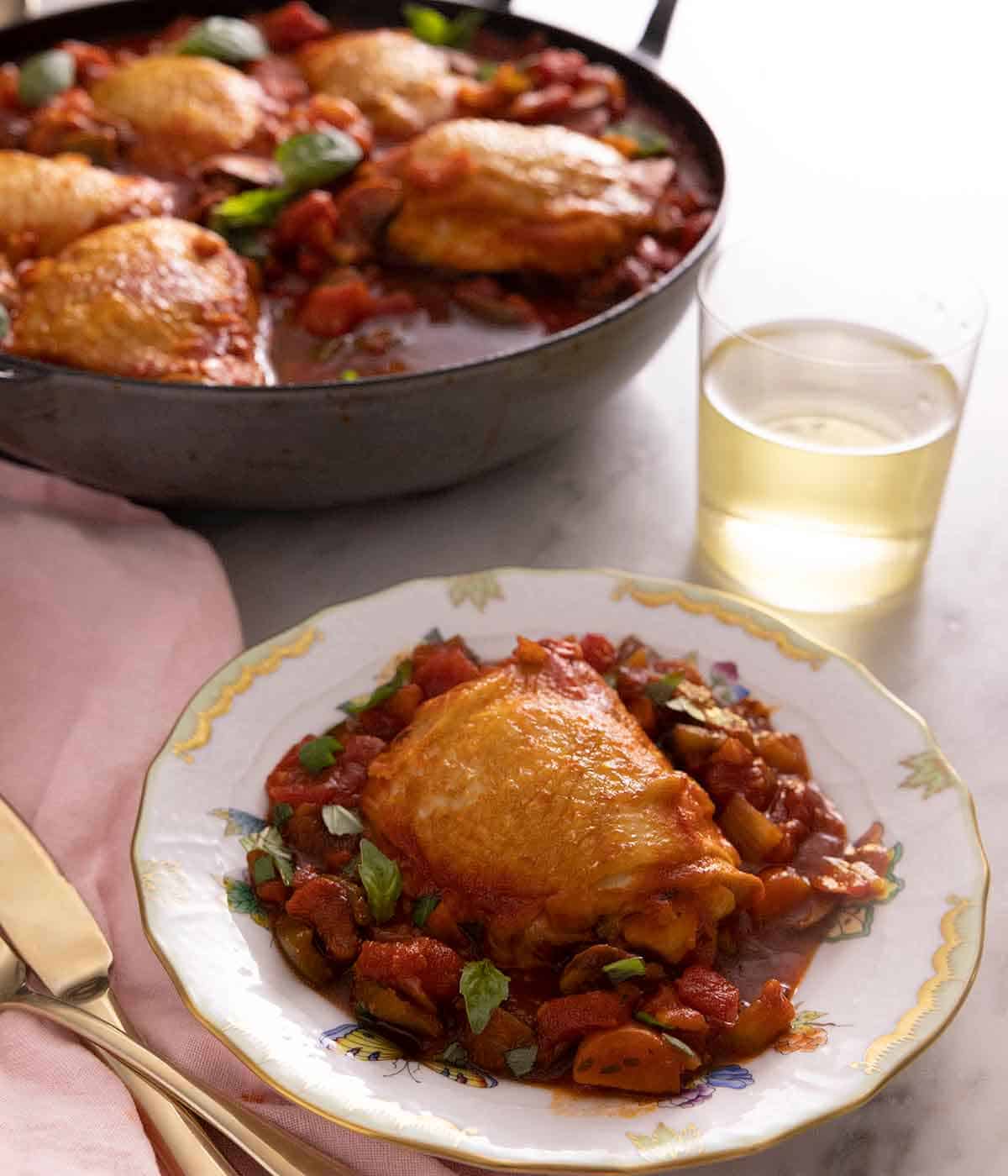 A plate with a serving of chicken cacciatore in front of a glass of wine and a pan with the rest of the food.