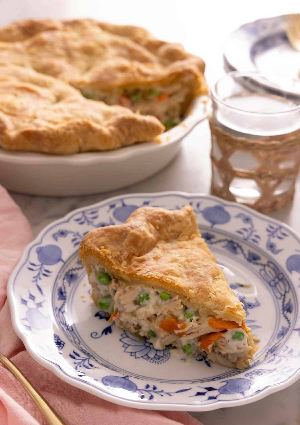 A plate of with a slice of chicken pot pie in front of a glass of water and the pie dish.