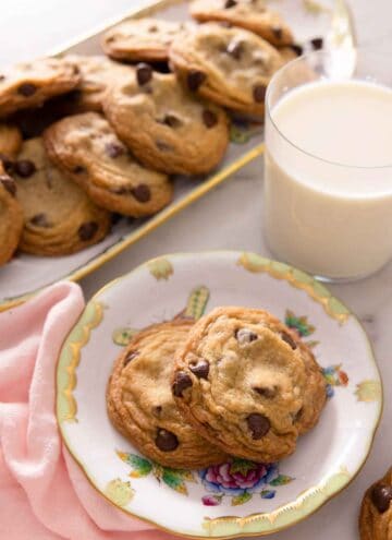 A plate with two chocolate chip cookies in front of a cup of milk and platter of cookies.