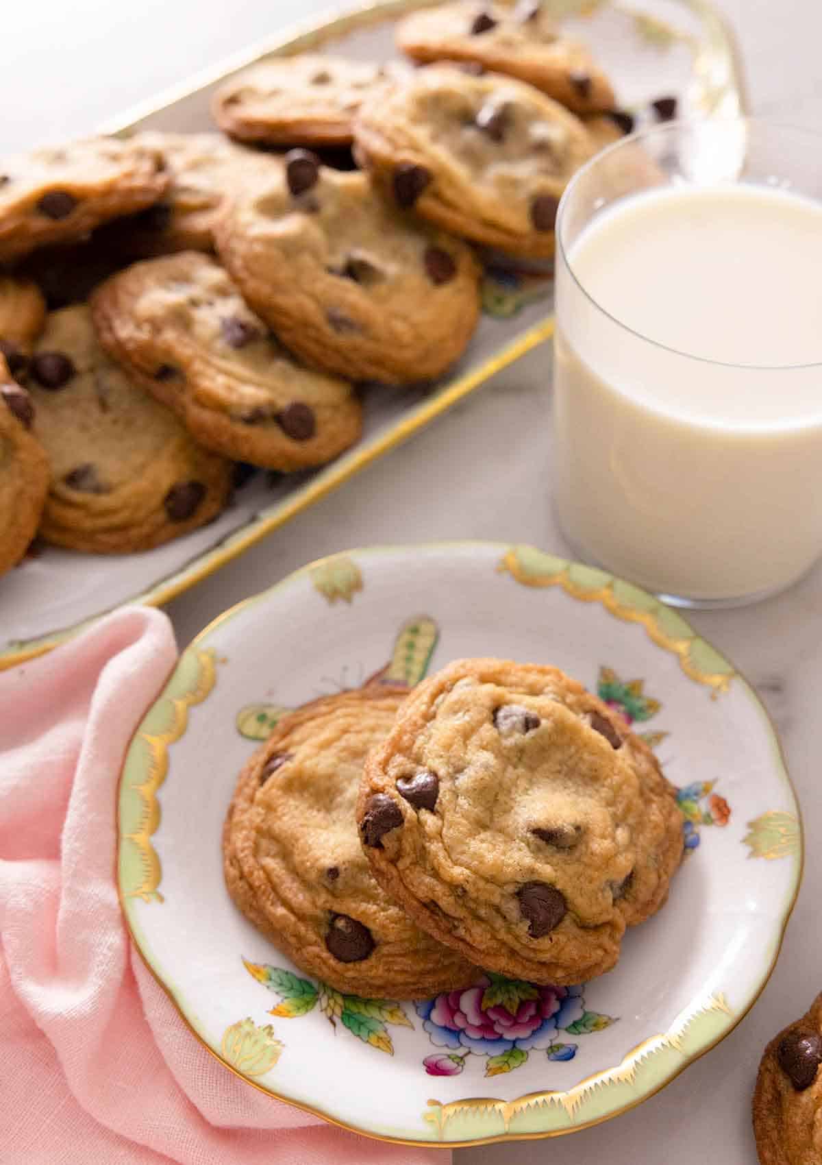 A plate with two chocolate chip cookies in front of a cup of milk and platter of cookies.
