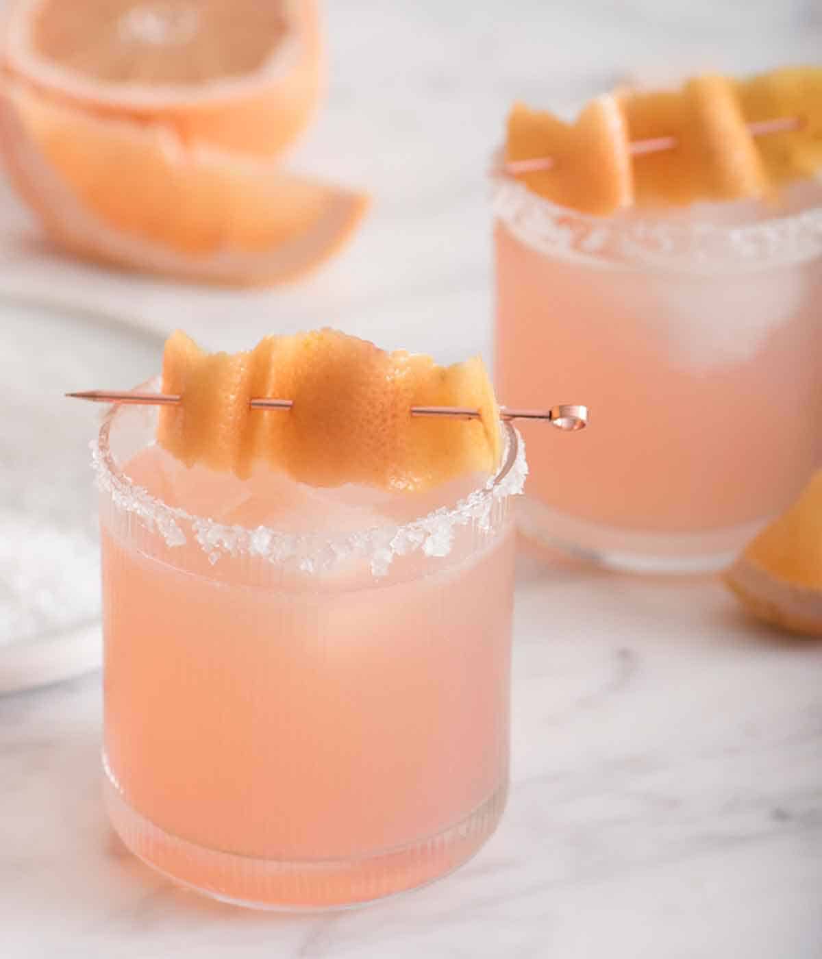 Close up of one of two paloma cocktails with salted rims and a grapefruit peel garnish.