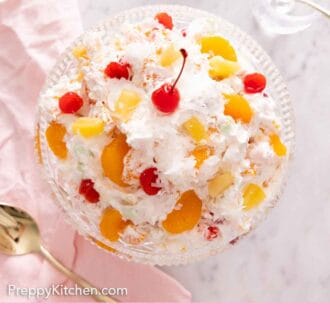 Pinterest graphic of the overhead view of a large bowl of ambrosia salad with a cherry on top beside a pink napkin and gold spoon.