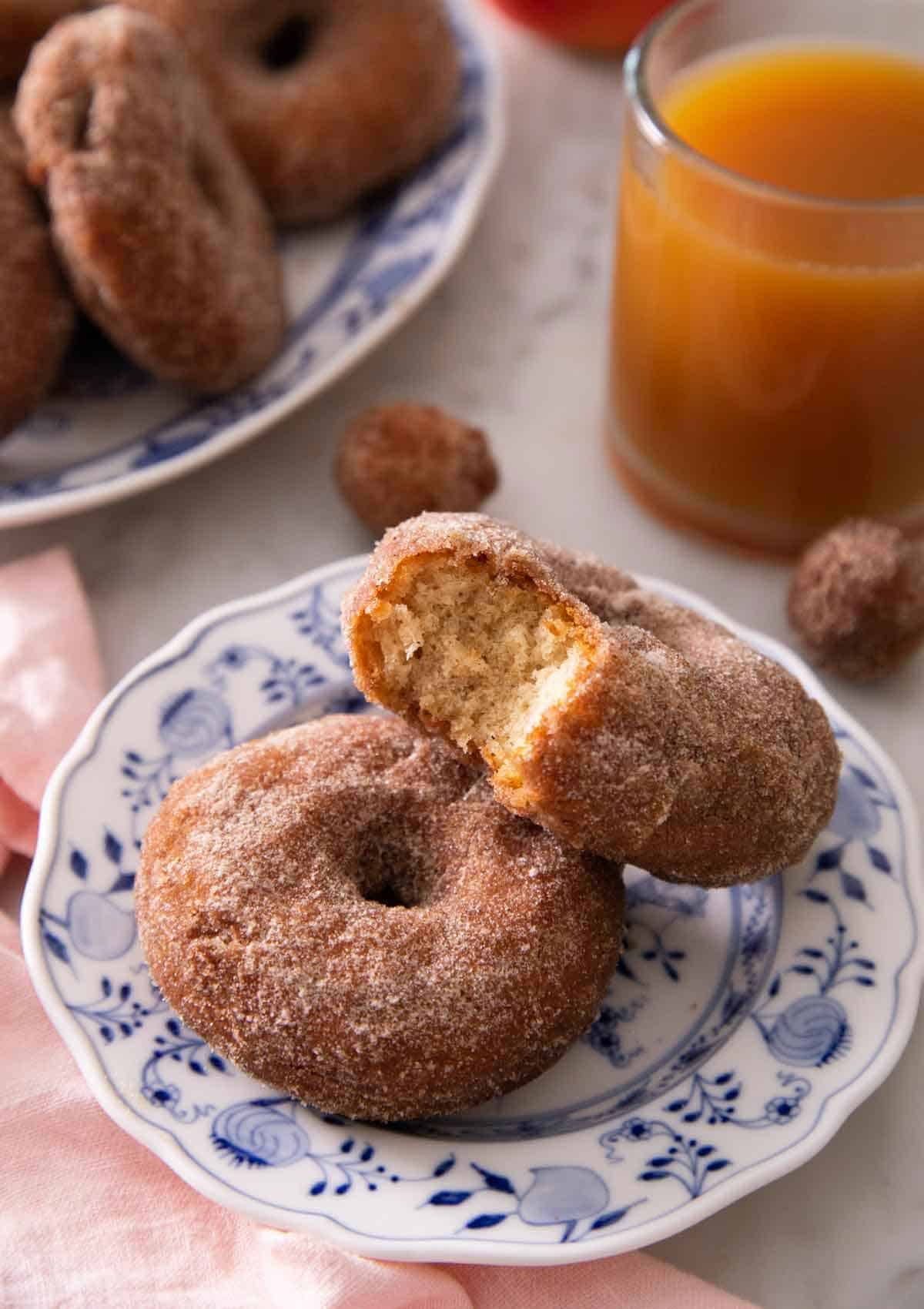Two apple cider donuts, with one with a bite taken out of it, on a plate in front of a cup of apple cider.