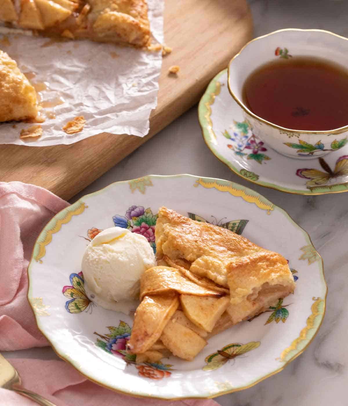 A plate with a slice of apple galette with a scoop of ice cream beside it in front of a cup of tea.