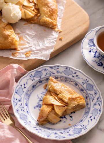 A plate with a slice of apple galette with a cutting board in the background with the rest of the galette along with a cup of tea.