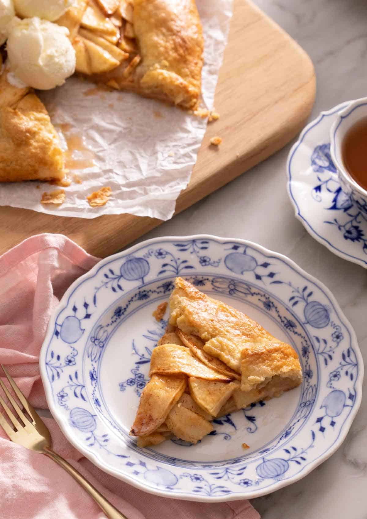 A plate with a slice of apple galette with a cutting board in the background with the rest of the galette along with a cup of tea.