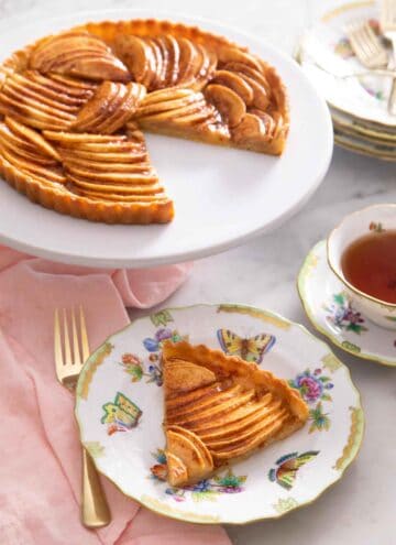 A serving of apple tart in a plate in front of the entire tart that it was cut out of.
