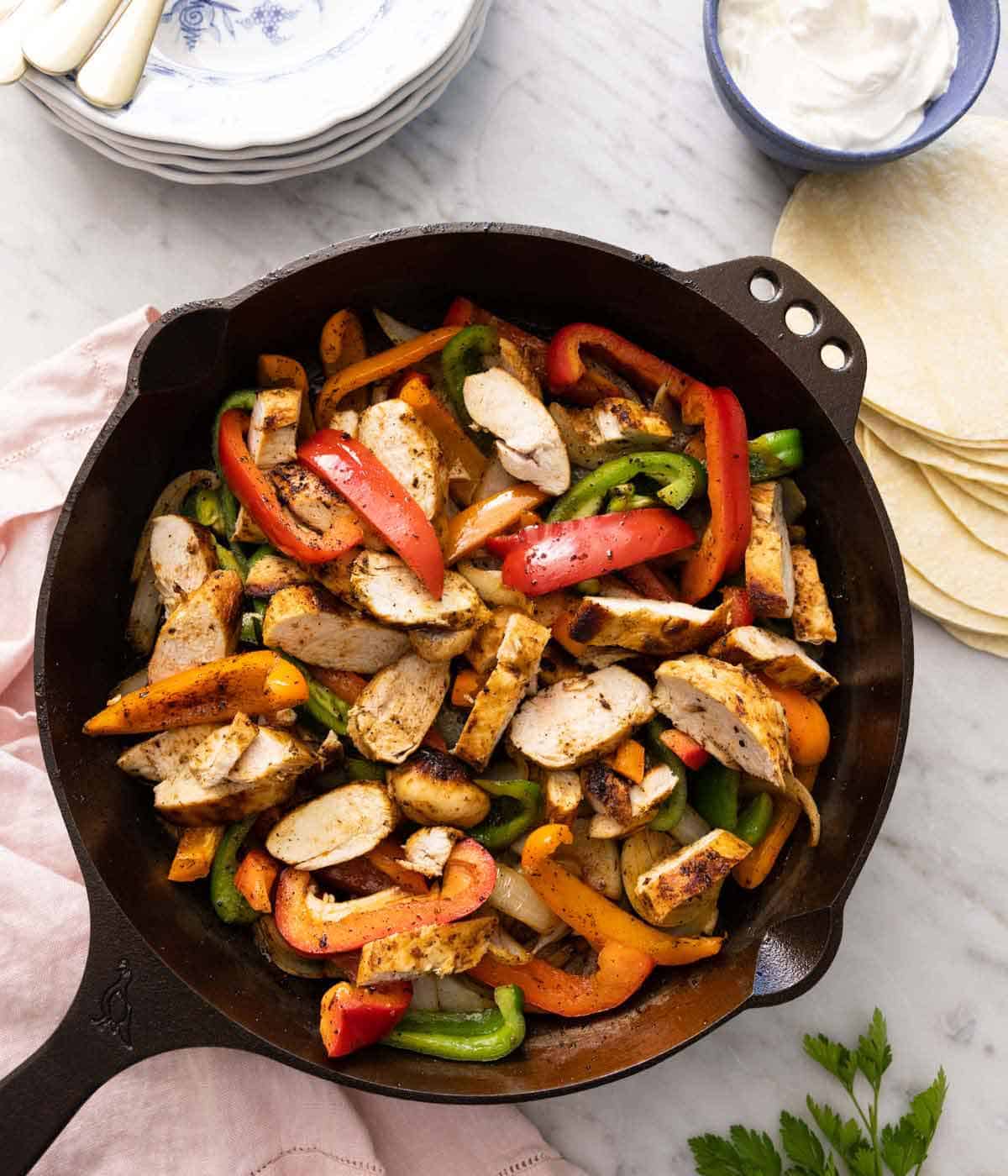 A cast iron skillet with chicken fajitas beside tortillas, sour cream, and a pink linen napkin.