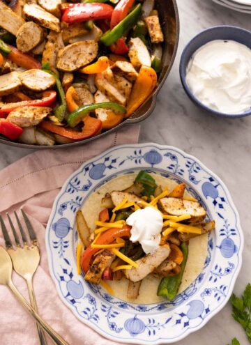 A plate with chicken fajitas on a tortilla in front of a pan of fajitas.