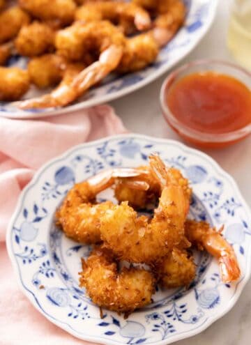 A small plate with five coconut shrimps in front of a dipping sauce and a platter with more shrimp.