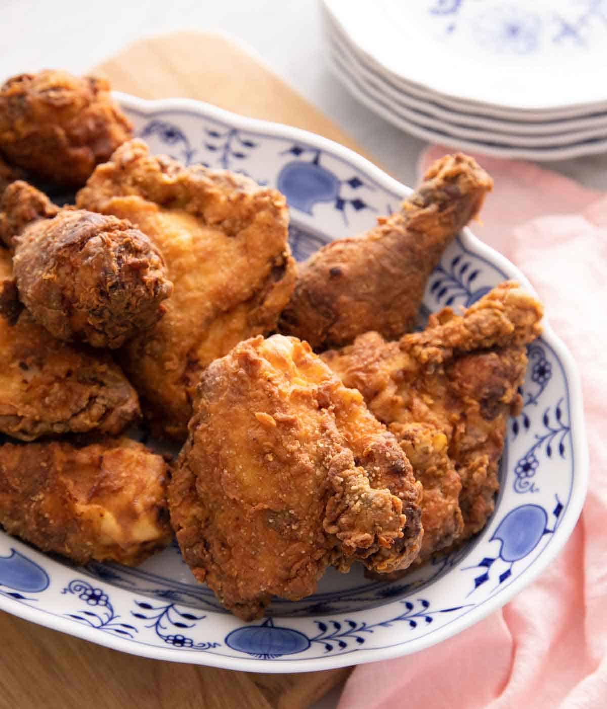 A serving platter with multiple pieces of fried chicken with a stack of plates in the background.