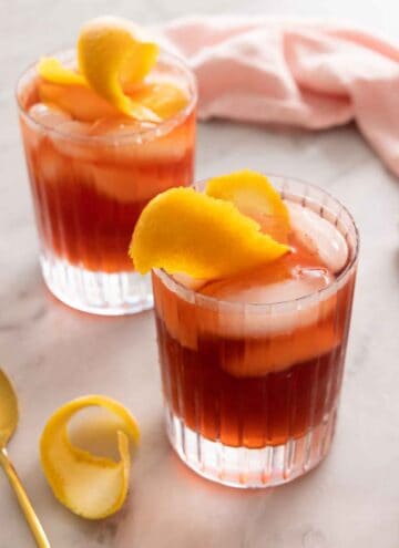 Two glasses of Negroni cocktails with orange peels as garnish.