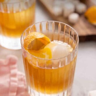 Pinterest graphic of a glass of old fashioned with an orange peel and large ice cube inside. A second one out of focus in the background.