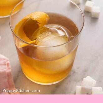Pinterest graphic a glass of old fashioned with a circular ice cube and orange peel inside and sugar cubes scattered around.