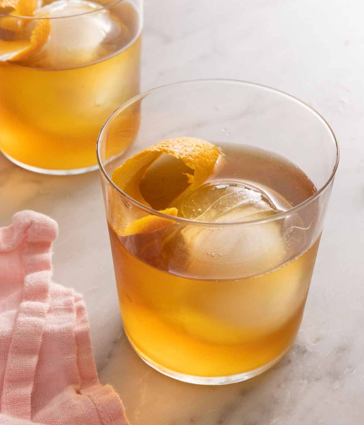 A glass of old fashioned with a spherical ice cube and orange peel inside. A second drink in the background.