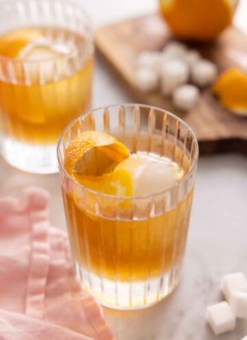 Two glasses of old fashioned with the ingredients in the background, out of focus.