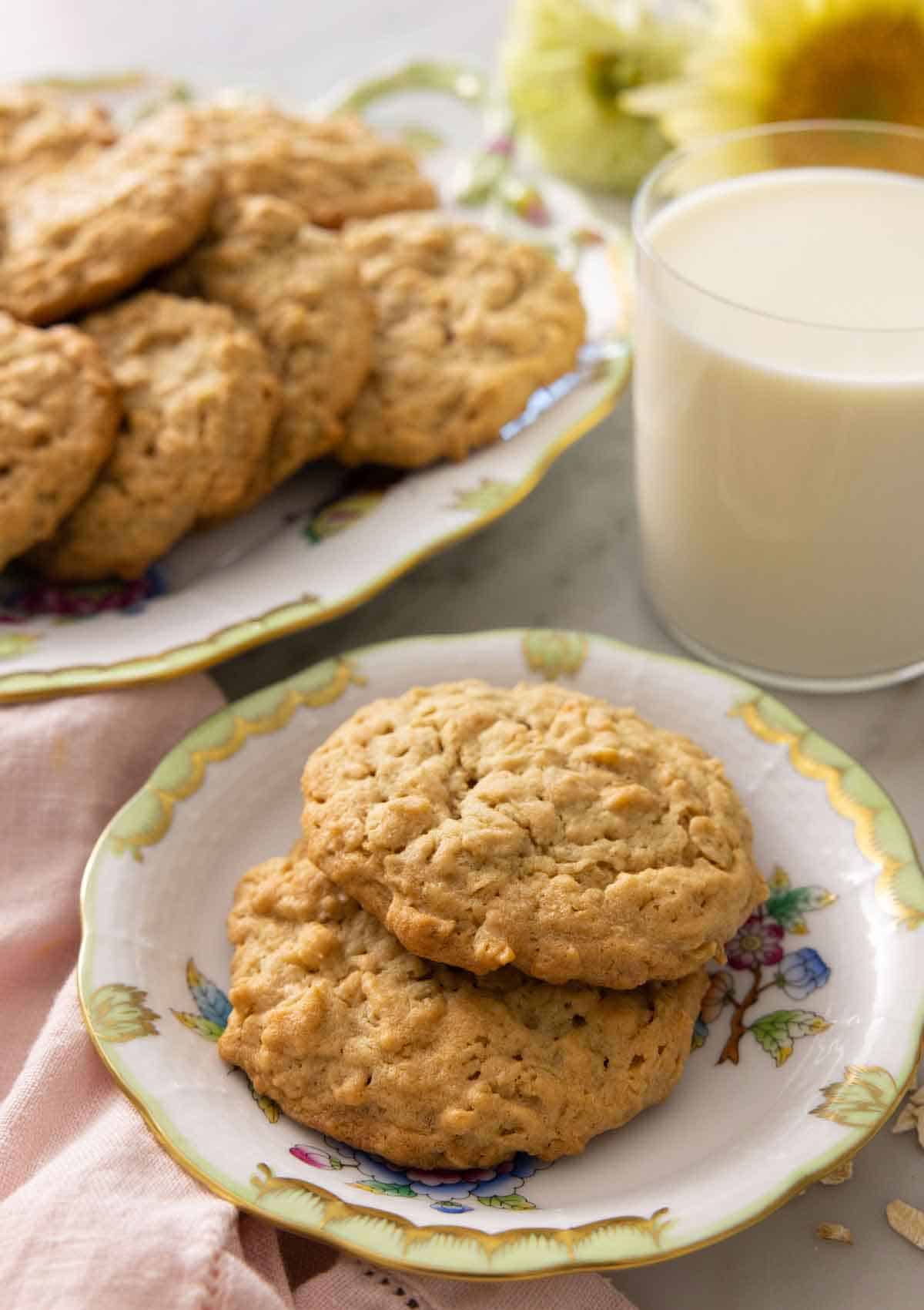 A plate with two peanut butter oatmeal cookies in front of a glass of milk and a platter of more cookies.