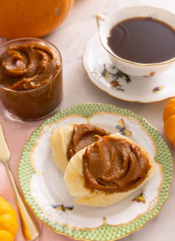 Two pieces of bread with pumpkin butter spread on top, in front of a glass of pumpkin butter and a cup of coffee.