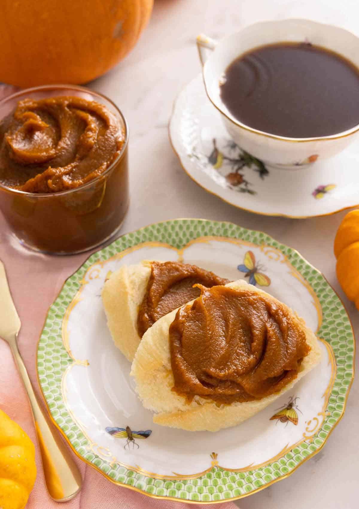 Two pieces of bread with pumpkin butter spread on top, in front of a glass of pumpkin butter and a cup of coffee.
