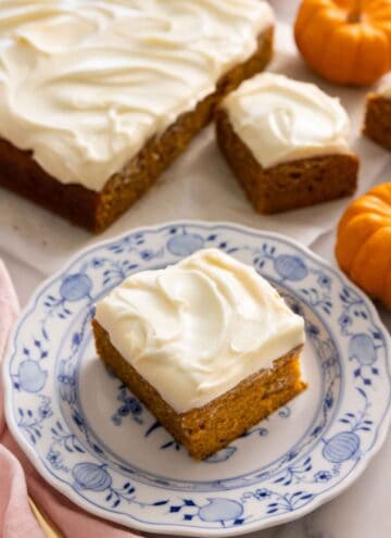 A square slice of pumpkin cake on a plate with the rest of the cake in the background.