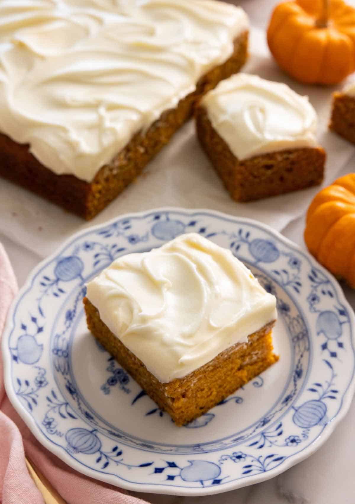 A square slice of pumpkin cake on a plate with the rest of the cake in the background.