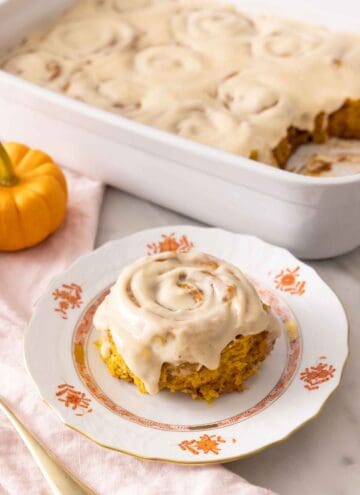 A plate with a single pumpkin cinnamon roll with cream cheese frosting on top in front of a baking pan with the rest.