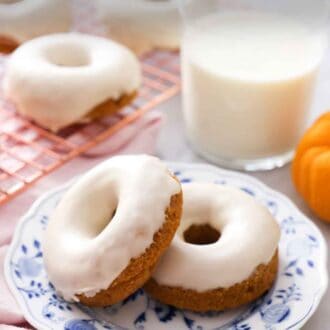 Pinterest graphic of two glazed pumpkin donuts with one propped up against the other in front of a cup of milk and a rack with donuts in the back.