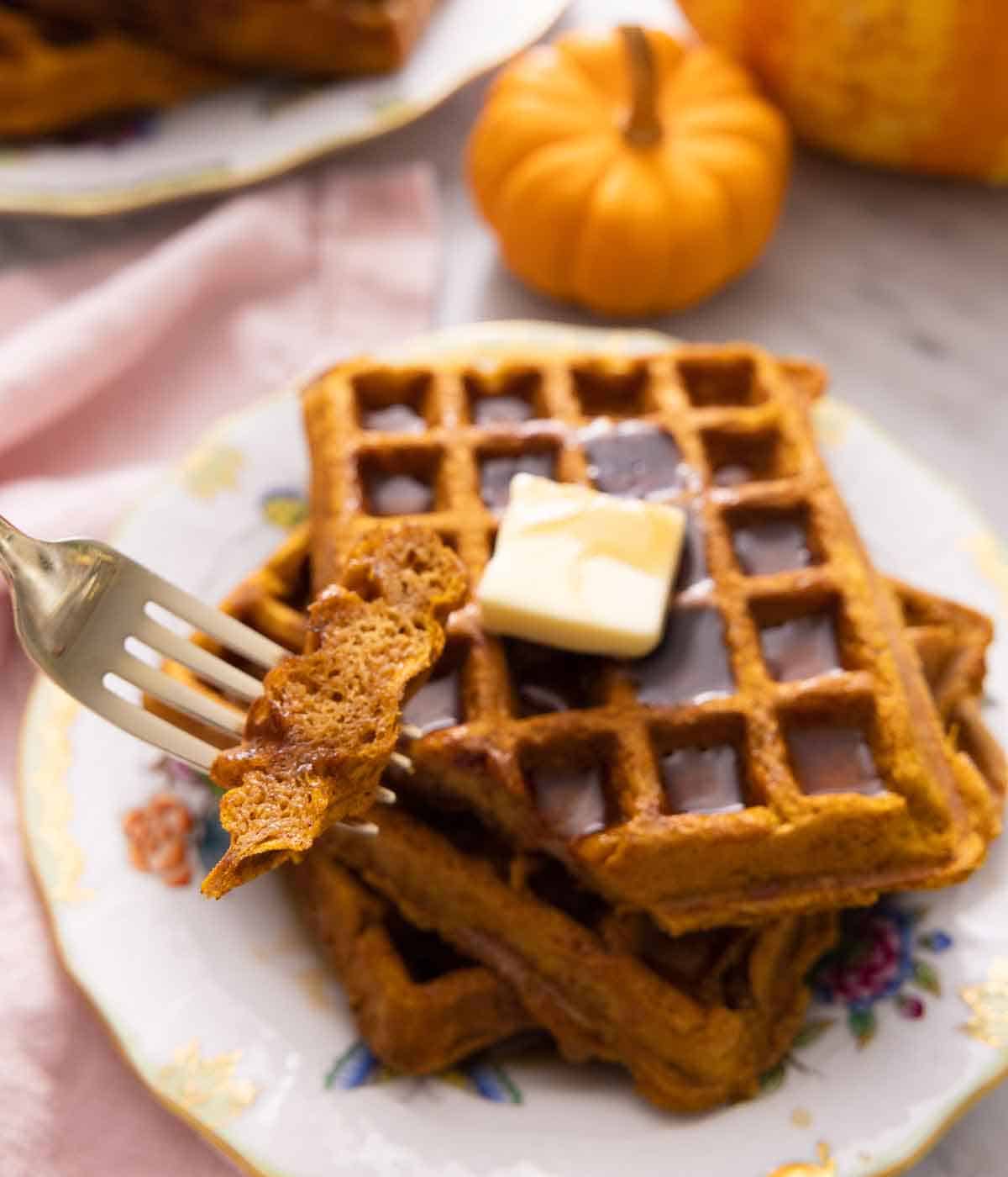 A fork with a piece of pumpkin waffle in front of a plate of waffles with syrup and butter.