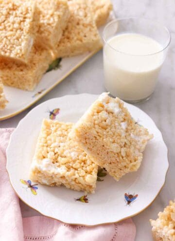 A plate with two squares of rice crispy treats in front of a glass of milk and a platter with more in the back.
