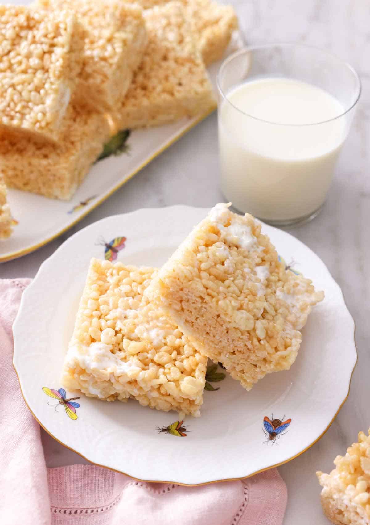 A plate with two squares of rice crispy treats in front of a glass of milk and a platter with more in the back.