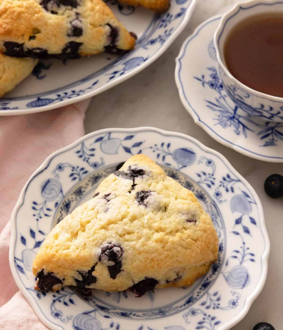 A plate with a blueberry scone in front of a cup of tea.