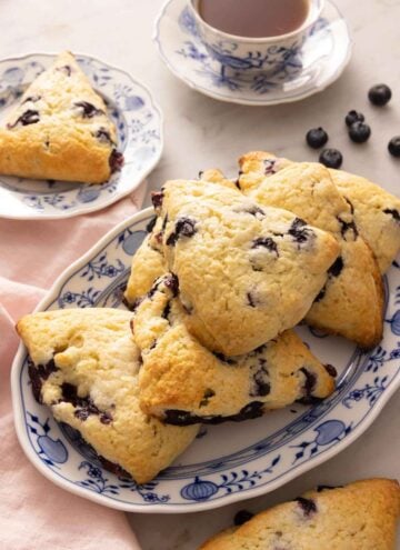An oval platter of blueberry scones in front of a cup of tea and scattered blueberries on the counter.