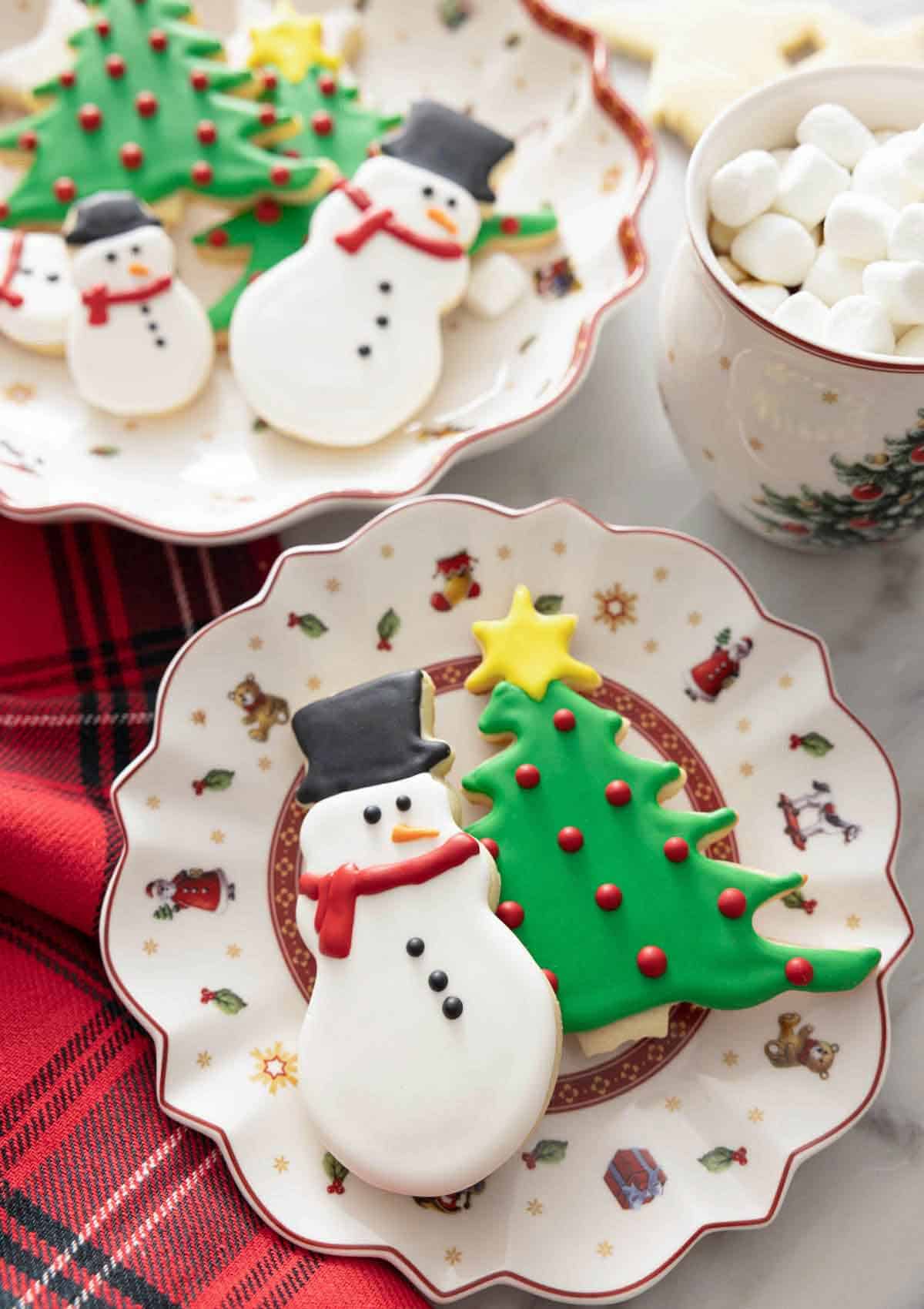 A snowman and Christmas tree cookie on a plate decorated with royal icing.